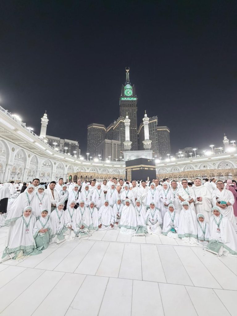 Rombongan besar jemaah Safari Suci, berpakaian ihram putih dan hijab putih, berpose untuk foto bersama di pelataran Masjidil Haram, Makkah pada malam hari, dengan Ka'bah terlihat jelas dan menara-menara masjid yang diterangi lampu di latar belakang, menjelaskan umrah berkunjung ke Baitullah