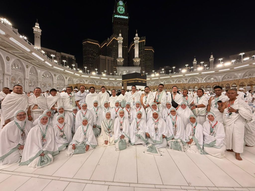 Rombongan jemaah Umrah Safari Suci berpakaian Ihram putih berfoto bersama di depan Ka'bah, Masjidil Haram Makkah pada malam hari, memahami makna umrah secara bahasa artinya kunjungan ke Baitullah